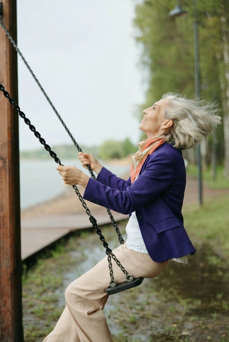 Older woman in a purple jacket on a swing outdoors, hair blowing, face lifted — representing vitality and strong bone health supported by MagZorb K2 + D3